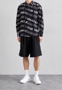 Svart skjorta med vitt textmönster, svarta shorts och vita sneakers. Skjortan har krage och knapplåsning fram. Släta texturer.