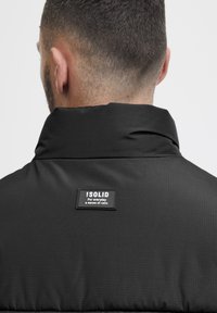 Black jacket with a high collar, smooth texture, and a rectangular brand label on the back featuring white text.