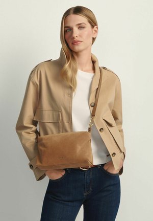 Kobieta o długich blond włosach ma na sobie beżową kurtkę, białą koszulę, ciemne dżinsy i nosi brązową zamszową torbę na ramię, trzymając ręce w kieszeniach.