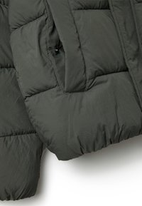 Giacca imbottita verde scuro con una texture liscia, dotata di tasca e design trapuntato. Il tessuto sembra leggero ma caldo.