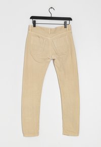 Pantalon beige taille slim avec une texture lisse, cinq poches et une taille mi-haute, suspendu à un cintre en plastique noir sur un fond blanc.