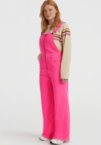Rødhåret person iført lysebrune overalls, beige langermet skjorte med striper, og hvite joggesko, står med hendene i lommene.
