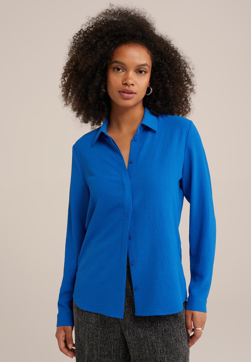 Camicia blu con bottoni e maniche lunghe, realizzata in un tessuto liscio, con colletto e una vestibilità ampia, abbinata a pantaloni scuri e testurizzati.