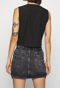 Person som bär en ärmlös svart topp och en urtvättad svart denimkort kjol med två bakfickor, som visar tatueringar på båda armarna.