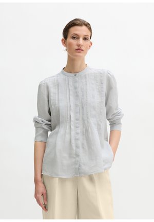 Vrouw die een lichtgrijs blouse met lange mouwen en verticale plooien draagt, gecombineerd met beige wijde broekspijpen, staand met één hand in haar zak.