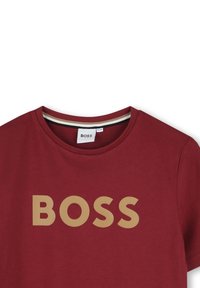 Camiseta de algodón roja con cuello redondo y mangas cortas, con un destacado logo "BOSS" beige impreso en la parte delantera.