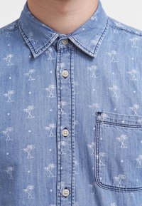 Camicia in denim azzurro chiaro con motivo di palme bianche e pois, colletto button-down e tasca sul petto con accenti cuciti.