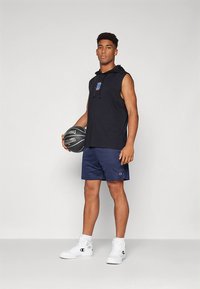 Melns bezroku kapuces džemperis ar logotipu, kopā ar tumši ziliem šortiem un baltām augstām kedām. Tur melnu basketbola bumbu, stāv vienkāršā fonā.