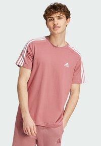 T-shirt a maniche corte in cotone rosa con design a tre strisce bianche sulle spalle e piccolo logo Adidas sul petto. Semplice scollatura a girocollo.