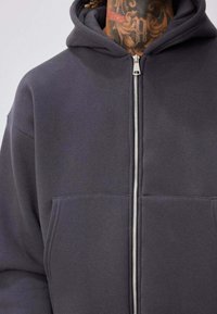 Sweat à capuche gris zippé avec capuche, doté de deux poches avant et d'une texture lisse. La fermeture éclair s'étend depuis l'encolure.