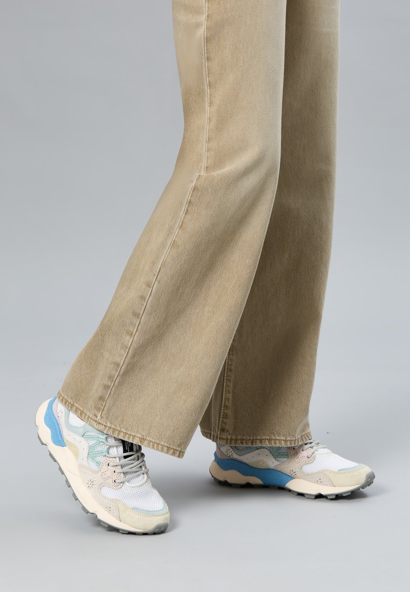 Person trägt beige, weit geschnittene Hosen und weiße Turnschuhe mit blauen und beigen Akzenten auf einem grauen Boden.