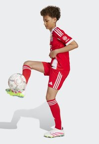 Rød fotballdrakt med hvite detaljer, bestående av en trøye og shorts, stripete sokker, og hvite fotballsko med rosa og grønne detaljer. Ball ved foten.