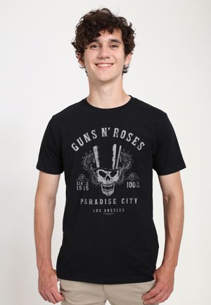 Jeune homme aux cheveux bouclés souriant, portant un t-shirt noir du groupe Guns N' Roses avec un motif de crâne et haut de forme.