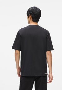 Čierne bavlnené tričko s krátkym rukávom a klasickým výstrihom, s rovnom okrajom a minimálnym brandovaním na spodnej časti. Zobrazený zadný pohľad.