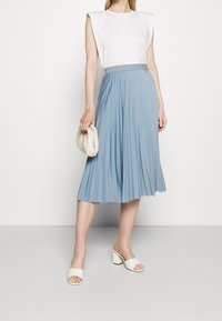 Hellblauer plissierter Midirock mit glitzerndem Bund, kombiniert mit einem weißen Kurzarmtop und weißen Sandalen mit Blockabsatz.