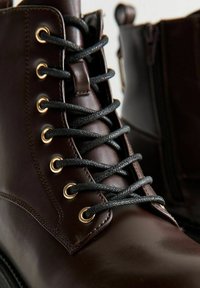 Bottes en cuir marron avec des lacets noirs et des œillets dorés. Elles présentent une texture lisse, un design à cheville haute et une fermeture éclair visible sur le côté.