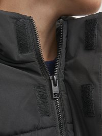 Black padded jacket featuring a front zipper, Velcro accents, and a high collar, showcasing smooth fabric and a structured design.