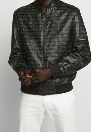 Homme portant une veste zippée noire ornée du texte répété "KARL LAGERFELD" et un pantalon blanc, les mains jointes devant lui.