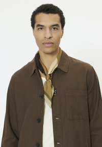 Veste marron à boutons avec une poche poitrine, portée sur une chemise claire. Une écharpe à motifs beige et marron est nouée autour du cou.