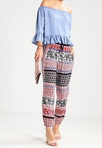 Top azzurro chiaro a spalla scoperta con maniche a volant, abbinato a pantaloni a fantasia e vestibilità morbida in rosso, nero e bianco. Clutch rosa pallido e sandali.