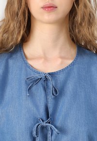 Denim top z okruglim izrezom, s dvema mašnama spredaj za zapiranje, v svetlo modri barvi z gladko teksturo.