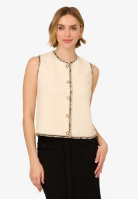 Krémová bezrukávová zkrácená vesta, s texturovanou tkaninou, černým lemem a zlatými knoflíky vpředu. Stylizována s černou sukní.