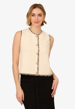 Veste cropped sans manches couleur crème, avec un tissu texturé, un liseré noir et des boutons dorés sur le devant. Portée avec une jupe noire.