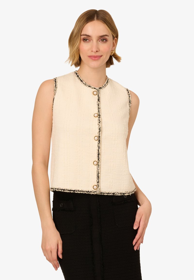 Krémová bezrukávová zkrácená vesta, s texturovanou tkaninou, černým lemem a zlatými knoflíky vpředu. Stylizována s černou sukní.