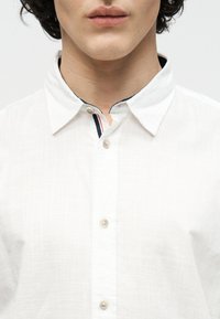 Κοντινό πλάνο ενός ατόμου που φοράει λευκό πουκάμισο με κουμπιά, με μικρή λεπτομέρεια από ναυτικό μπλε και κόκκινες ρίγες στο εσωτερικό του γιακά.