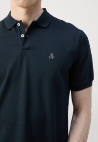 Granatowa koszulka polo z teksturowanej tkaniny, z dwoma guzikami i haftowanym logo na lewej piersi. Krótkie rękawy.