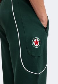 Κοντινή λήψη σκούρων πράσινων παντελονιών με λευκές ρίγες και σήμα της Red Star FC κοντά στην τσέπη όπου ένα χέρι είναι μερικώς εισαχθεί.