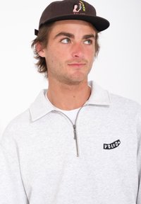 Sudadera gris claro con cremallera y cuello alto, con un logotipo negro de "VOLCOM". Combina con una gorra marrón oscura que presenta acentos gráficos coloridos.