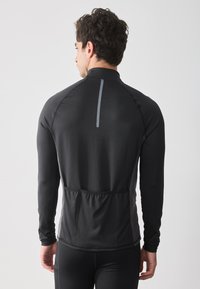 Camiseta deportiva de manga larga negra con un alto cuello, tejido texturizado y una franja vertical reflectante en la parte posterior. Cuenta con un diseño ajustado y un bolsillo trasero.