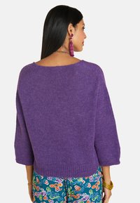 Lila gestrickter Pullover mit weit ausgeschnittenem Ausschnitt, drei Viertel-Ärmeln, geripptem Saum und weicher Textur. Bunte gemusterte Hosen sind darunter sichtbar.