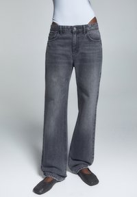 Modelo vistiendo jeans anchos de color negro desgastado con bolsillos frontales y cierre de botón, combinados con zapatos planos negros sobre un fondo gris.