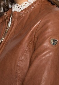 Hnědá kožená bomber bunda má zipový uzávěr, propletený detail límce a malý emblém na rukávu. Měkká textura a přiléhavý design.