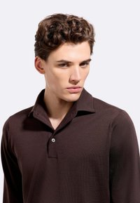 Polo a maniche lunghe marrone con pattina a bottoni e colletto classico. Realizzato in tessuto morbido, presenta una superficie liscia e un design aderente.