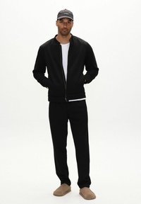 Svart zip-up jacka med ställbar krage, tillsammans med matchande svarta byxor. Modellen har på sig en vit skjorta och en mörk keps, med beige tofflor.