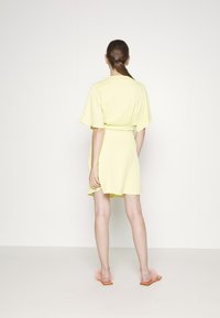 Vestido corto amarillo con mangas aladas y cintura ceñida con cinturón. Textura suave y hasta la rodilla. La modelo lleva sandalias de slide naranjas.