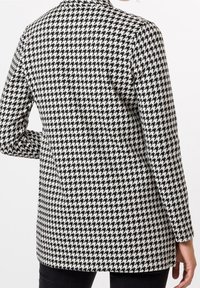 Mujer vistiendo un blazer de estampado de pata de gallo en blanco y negro, con mangas largas, de pie con una mano en la cadera.