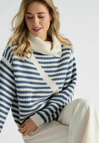 Gestreepte gebreide trui in marineblauw en crème, met een sjawlcollar met knoopdetails en een losse pasvorm, gecombineerd met lichte broek.