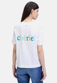 White cotton t-shirt, short sleeves, featuring multicolored text "chérie!" on the back, with a small green heart detail at the neck.