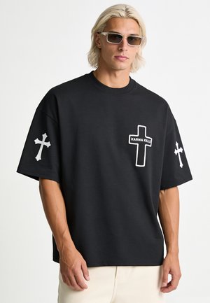Blonder Mann mit rechteckiger Sonnenbrille und schwarzem Hemd mit weißen Kreuzmustern, darunter eins mit der Aufschrift "KARMA KILLS" auf der Brust.