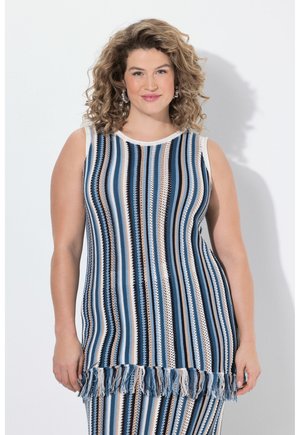Donna con capelli ricci che indossa un top in maglia senza maniche a righe verticali blu, bianche e beige con frange all'orlo, in piedi davanti a uno sfondo bianco.