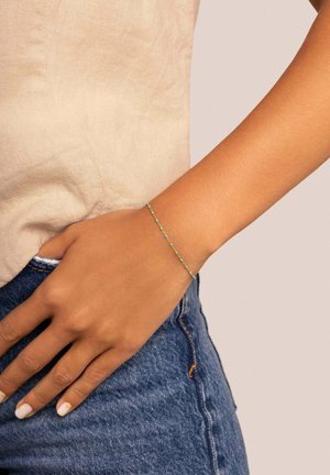 Bracciale dorato con piccole perle di turchese, design di catena delicato e un'estetica semplice e minimalista su uno sfondo color carne.