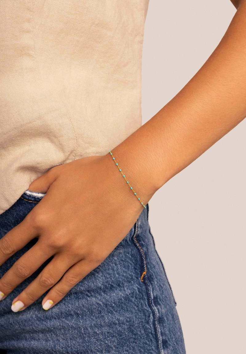 Pulsera de tono dorado con pequeñas cuentas de turquesa, diseño de cadena delicada y una estética simple y minimalista sobre un fondo en tonos de piel.
