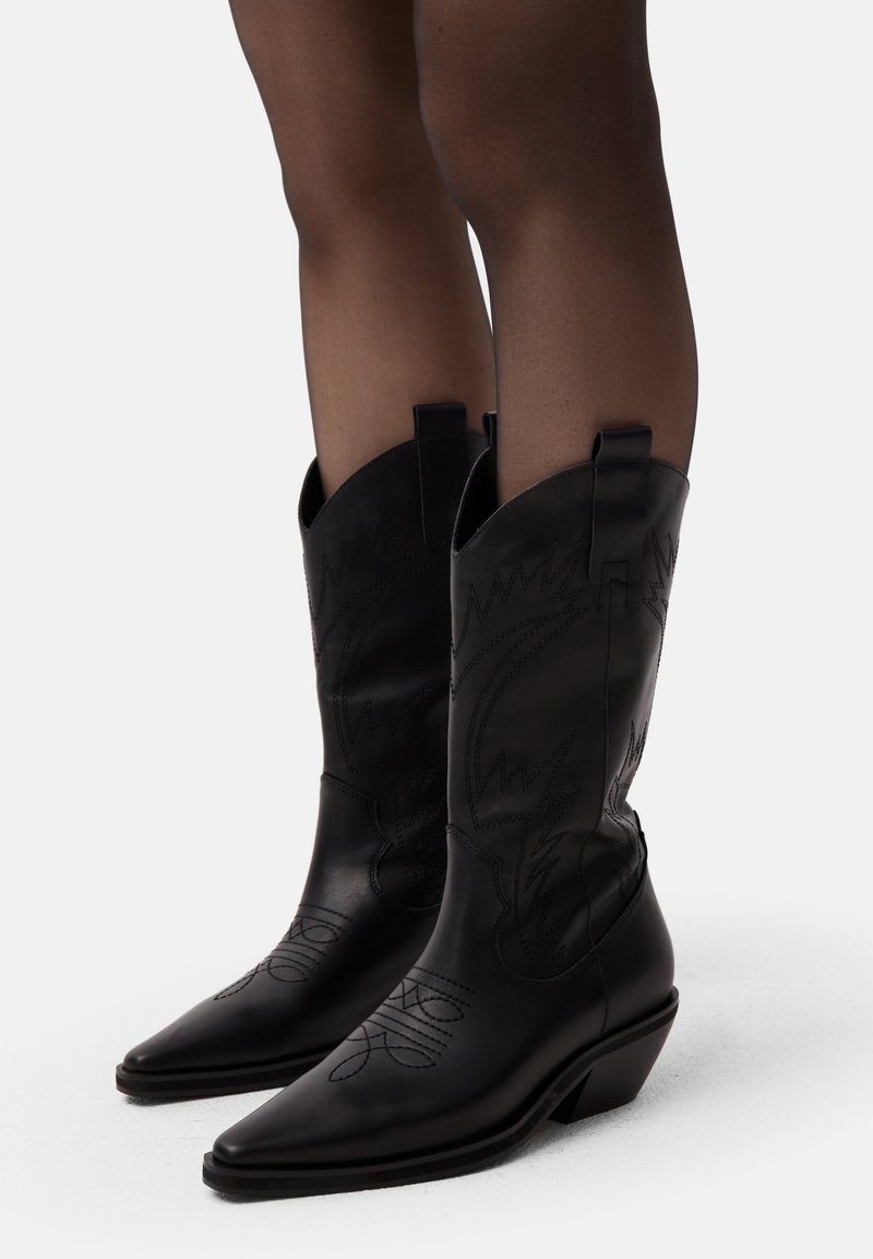 Botas de vaquero de cuero negro con punteras afiladas, costuras decorativas, lengüetas para tirar y un tacón bajo. Textura suave con patrones intrincados.