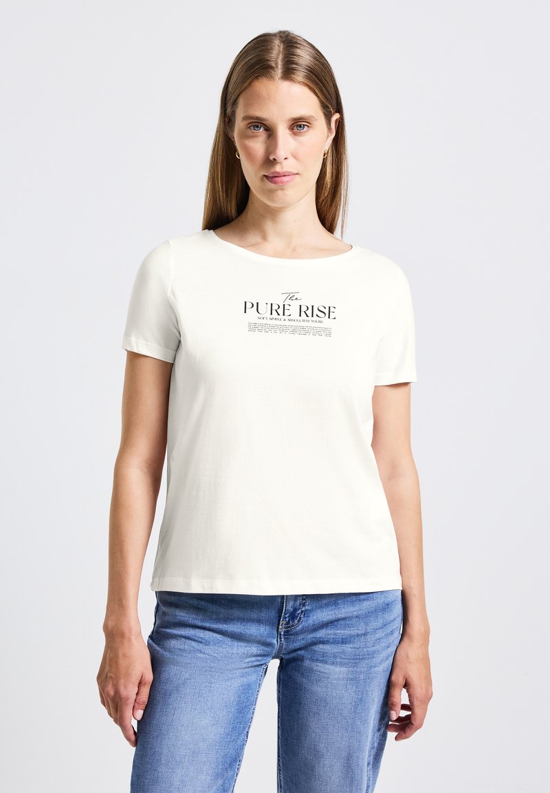 Nainen, jolla on pitkät hiukset, pukeutuneena valkoiseen lyhythihaiseen T-paitaan, jossa on teksti "The Pure Rise", sekä sinisiin farkkuihin tasaisella vaalealla taustalla.