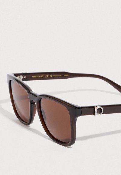 Occhiali da sole marrone scuro con montatura quadrata, lenti marroni e dettaglio hardware circolare argento sulle aste. Made in Italy.