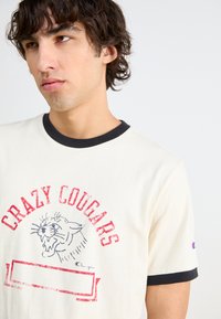 Tricou crem cu mânecă scurtă, guler și manșete negre, având inscripția roșie "CRAZY COUGARS" și un grafic cu fața unei pume.